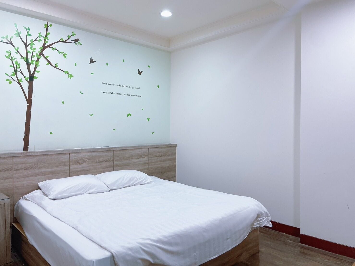 Een slaapkamer met een bed, nachtkastje, houten hoofdbord en een muur met een boomsticker en tekst.
