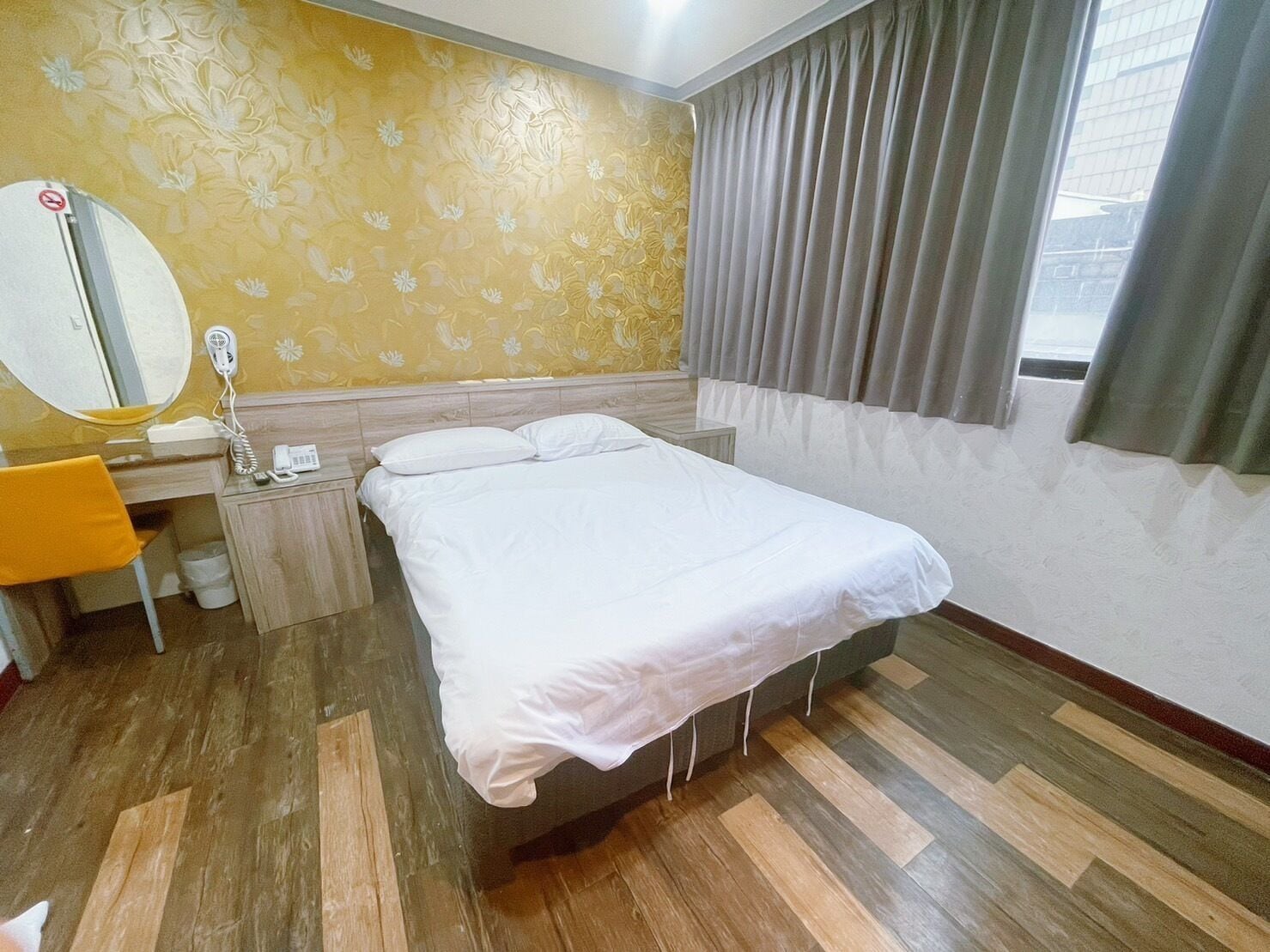 A hotel room with a bed, a desk with a telephone, a mirror, and a window with curtains.