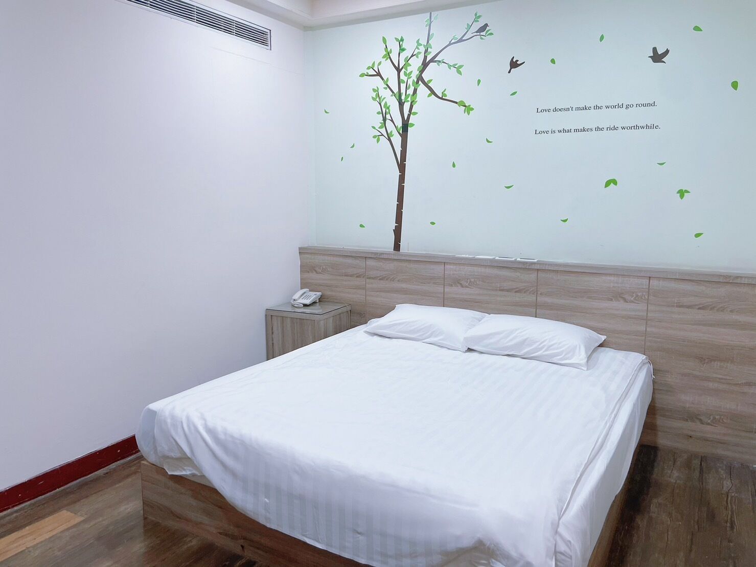 Una cama bien hecha en una habitación de hotel, con una obra de arte en la pared de un árbol y pájaros, y una mesita de noche con un teléfono.