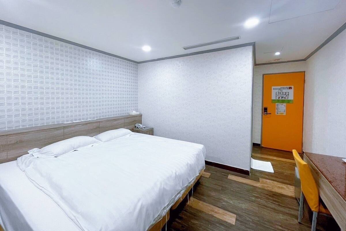 A hotel room with a bed, a wooden nightstand, a small table, and an orange door.
