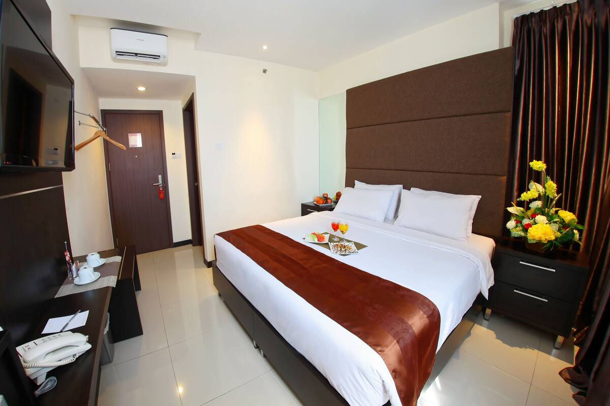 A hotel room with a large bed, a bedside table with a lamp, a TV mounted on the wall, and a bathroom door in the background.