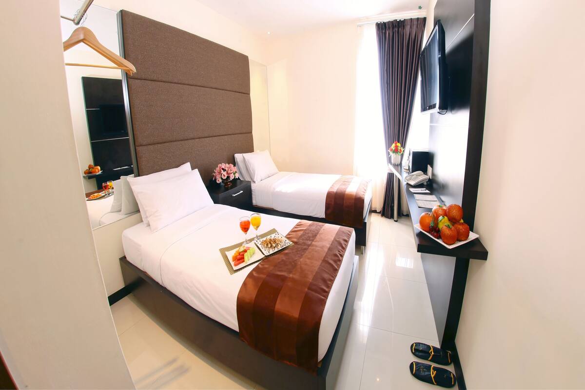 A hotel room with two beds, a flat-screen TV, a small table with fruit, and slippers on the floor.