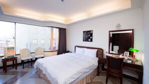 A hotel room with a large bed, a desk with a mirror, a chair, a desk lamp, and a view of the city through large windows. - Peach Hotel (Hsinchu)