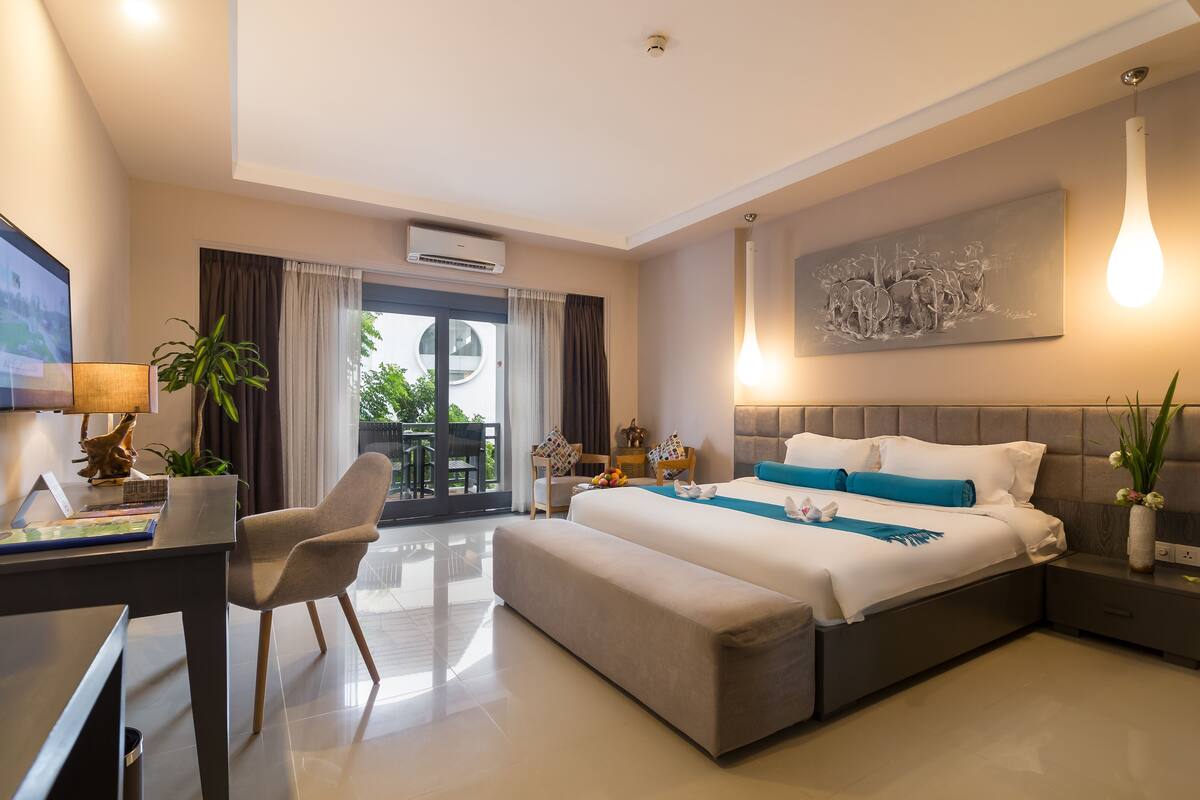 A modern hotel room with a large bed, a desk with a laptop, a chair, a bedside table, and a view of the outdoors.