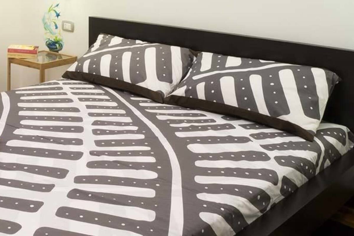 A neatly made bed with a patterned bedspread and pillows, a bedside table with a lamp and a small decorative item, and a tray with breakfast items on the bed.