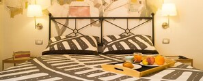 Una habitación de hotel con una cama que tiene un edredón estampado con motivos geométricos, dos lámparas de noche, una bandeja con frutas y una botella, y una pared con pintura descascarada.