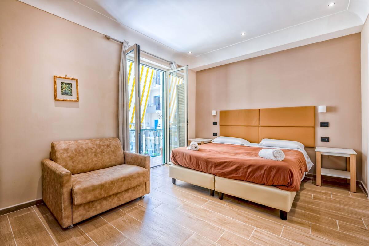 Een moderne hotelkamer met een bank, een tweepersoonsbed, een nachtkastje en uitzicht op buiten.