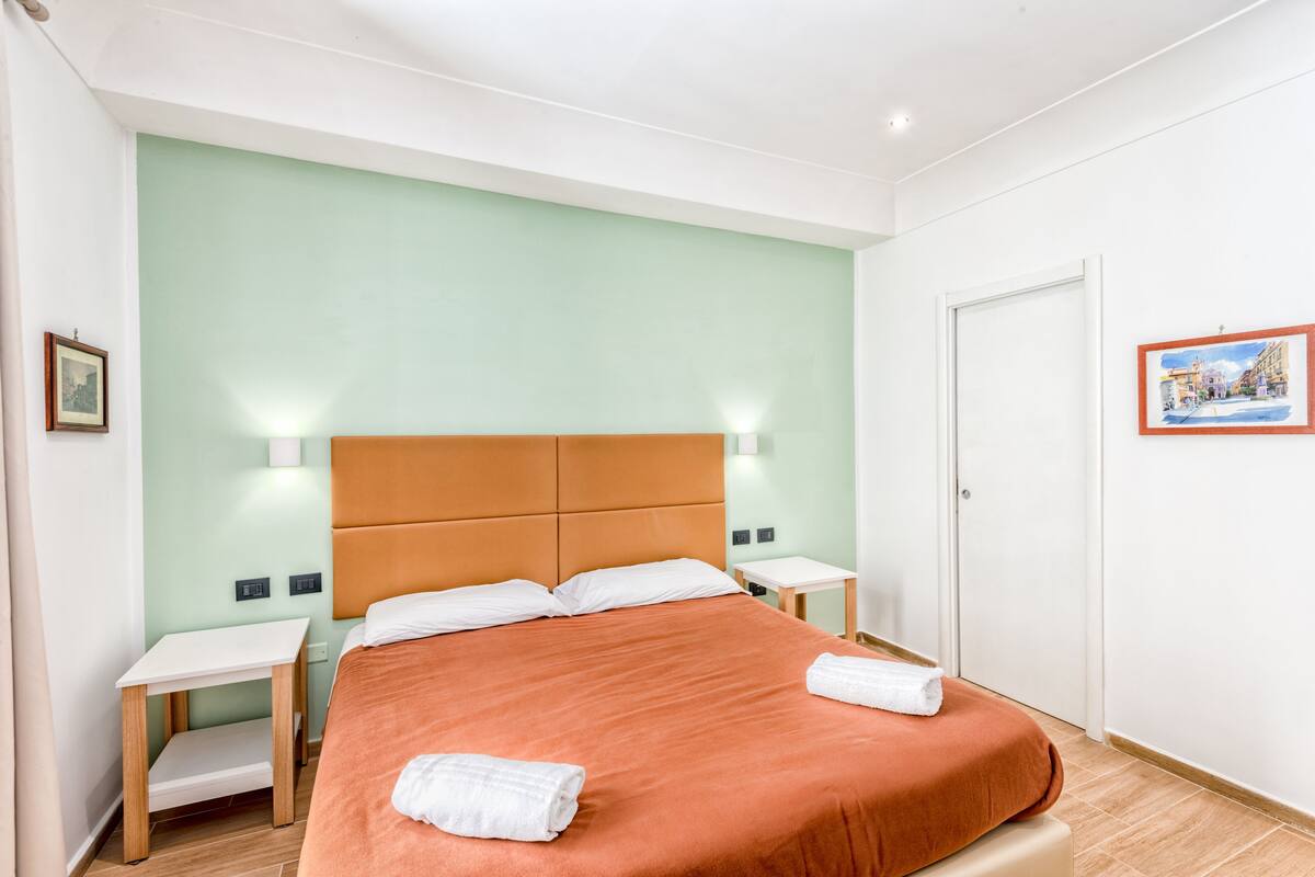 Een hotelkamer met een bed, nachtkastje, een klein tafeltje en een deur.
