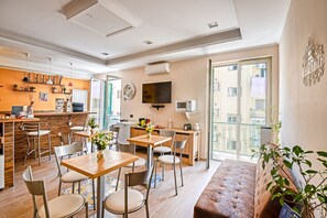A modern living room with a sofa, dining table, and chairs. There is a kitchen area with a counter and appliances. A TV is mounted on the wall, and there is a balcony with a view of the city. - Giacomino (Sorrento)