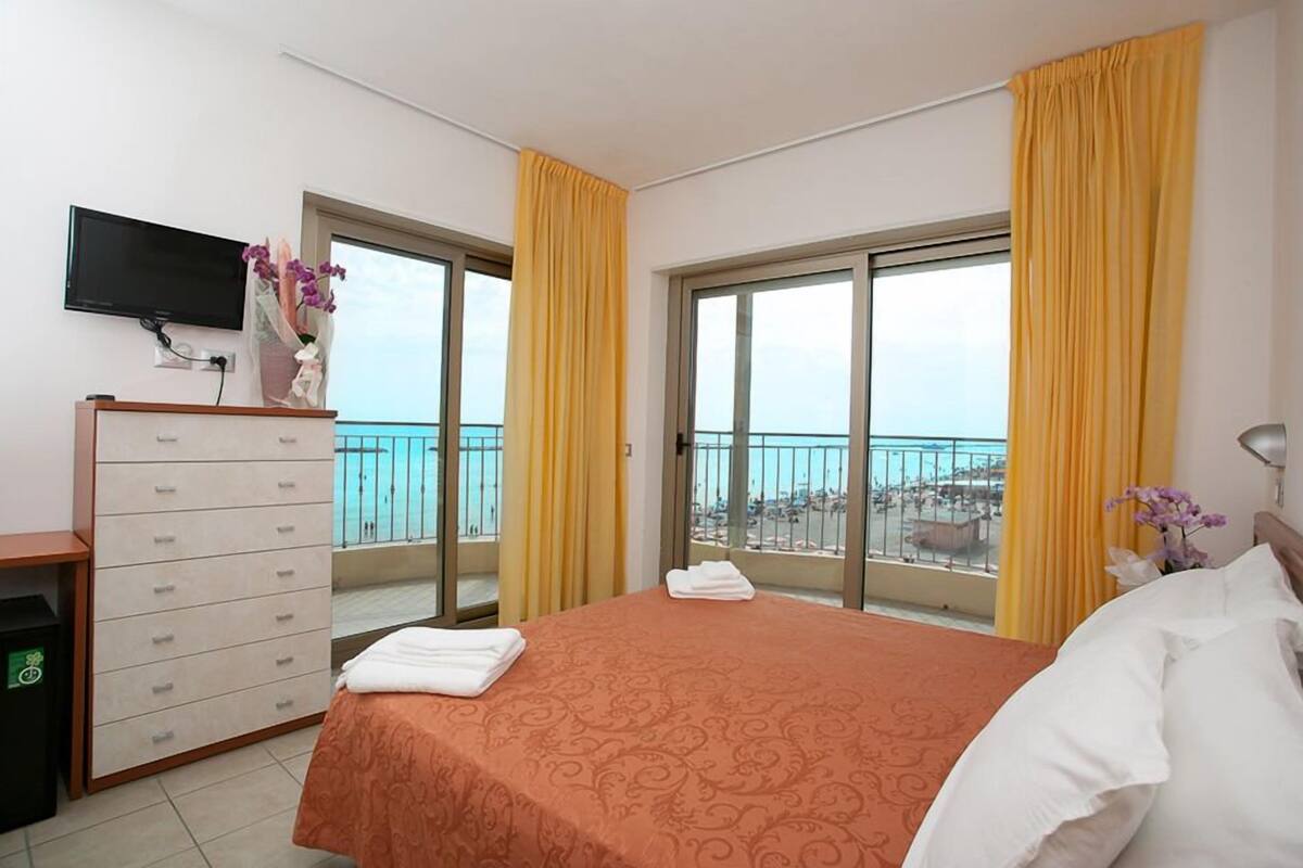 A hotel room with a bed, a dresser, a television, and a balcony with yellow curtains.
