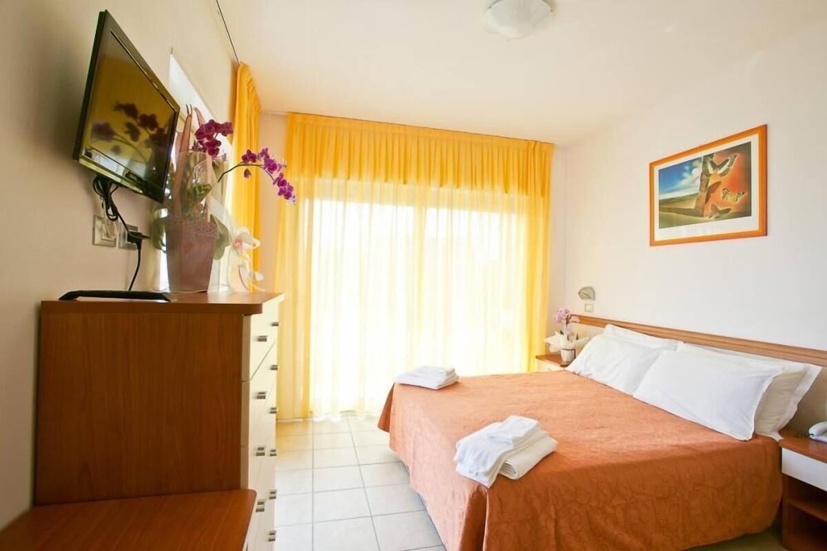 A hotel room with a bed, a desk with a computer, a TV, and a window with curtains.