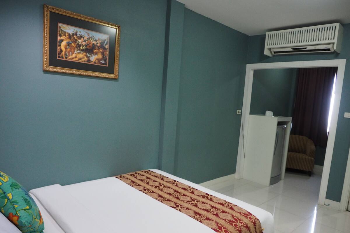 A hotel room with a bed, a framed picture on the wall, an air conditioning unit, and a door leading to another room.
