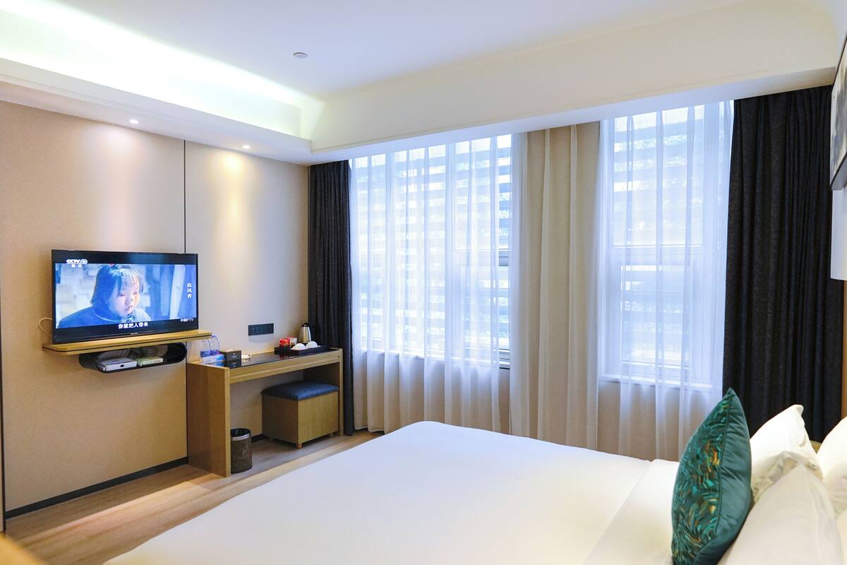 A hotel room with a bed, a TV mounted on the wall, a desk, and a window with curtains.