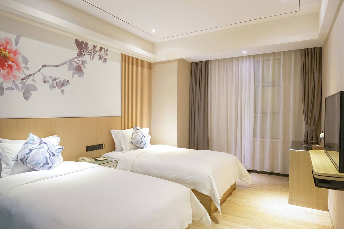 A hotel room with two beds, a wooden headboard, a large floral artwork on the wall, a desk with a mirror, and a window with curtains.
