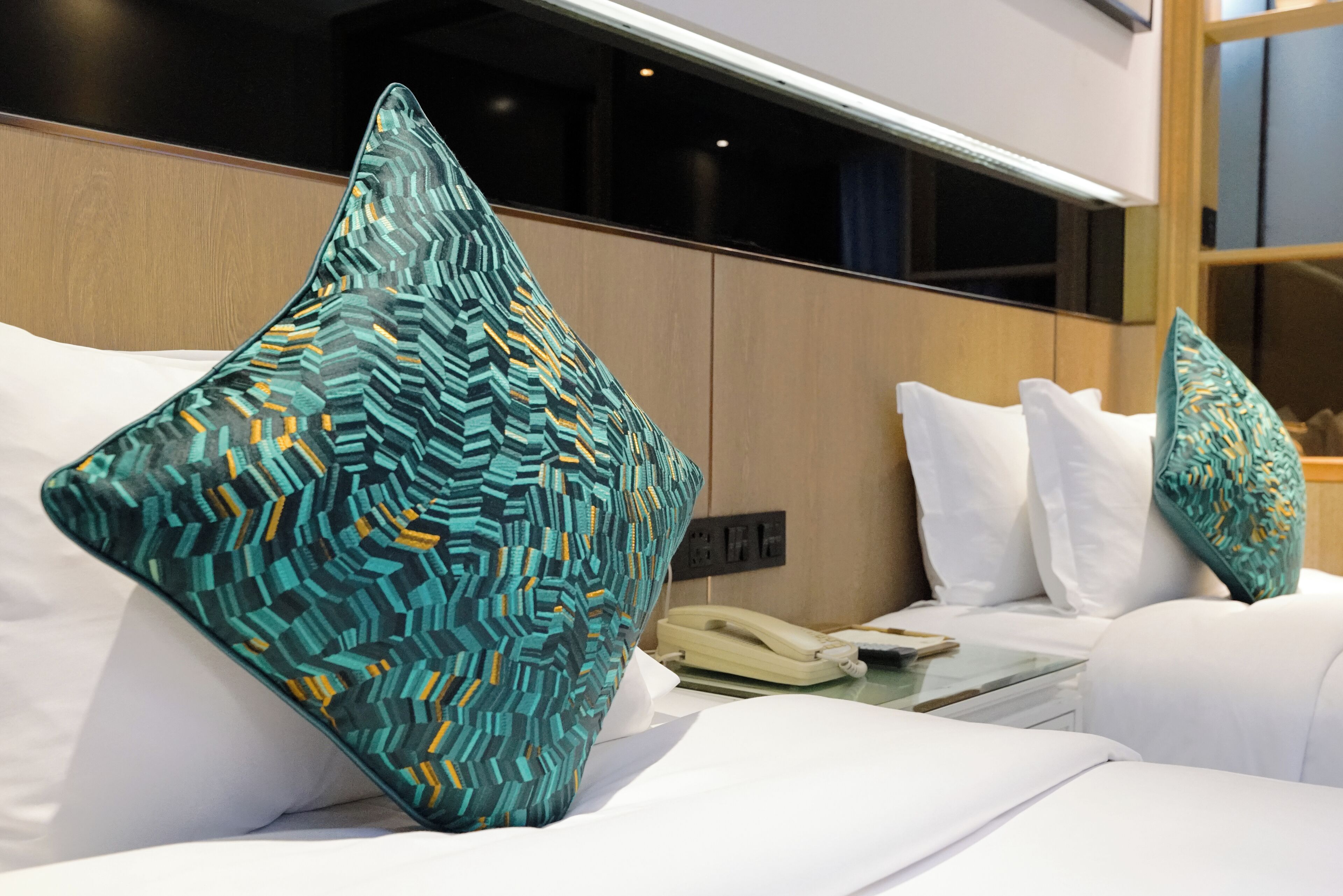 A hotel room with a bed, white pillows, a patterned pillow, a bedside table with a phone, and a window with blinds.