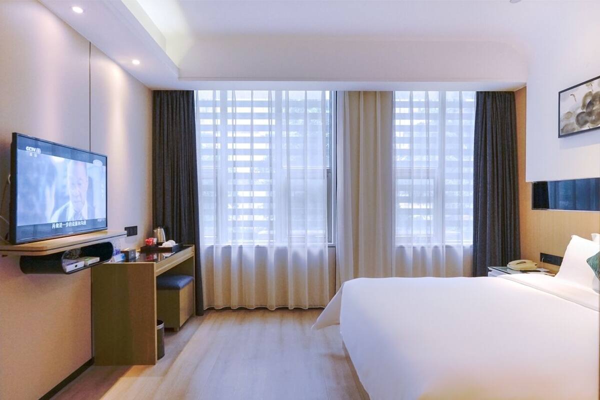 A hotel room with a bed, a desk with a television, a window with curtains, and a wall-mounted phone.
