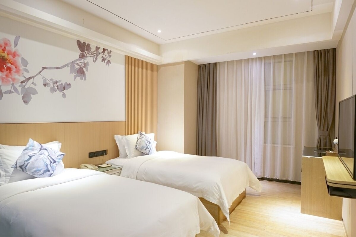 Hotelkamer met twee bedden, een houten hoofdbord, een schilderij aan de muur, een bureau met een televisie, en een raam met gordijnen.