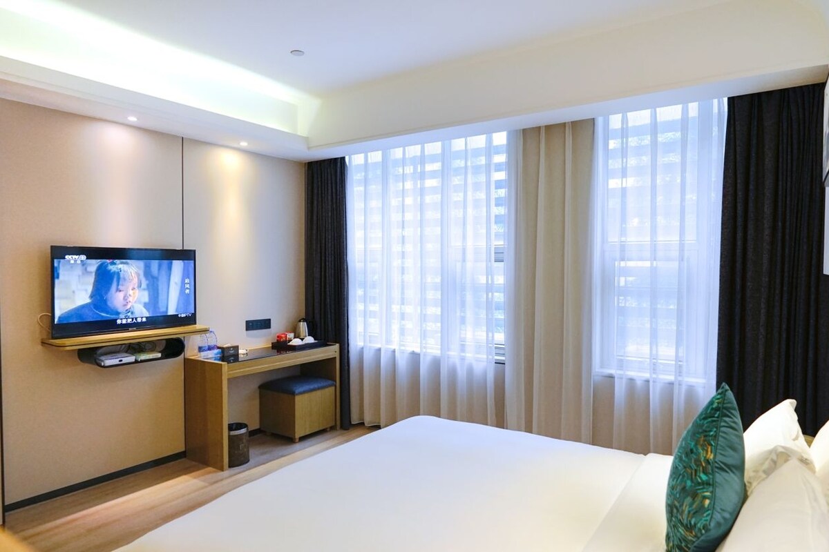 A hotel room with a bed, a desk, a television, and large windows with curtains.