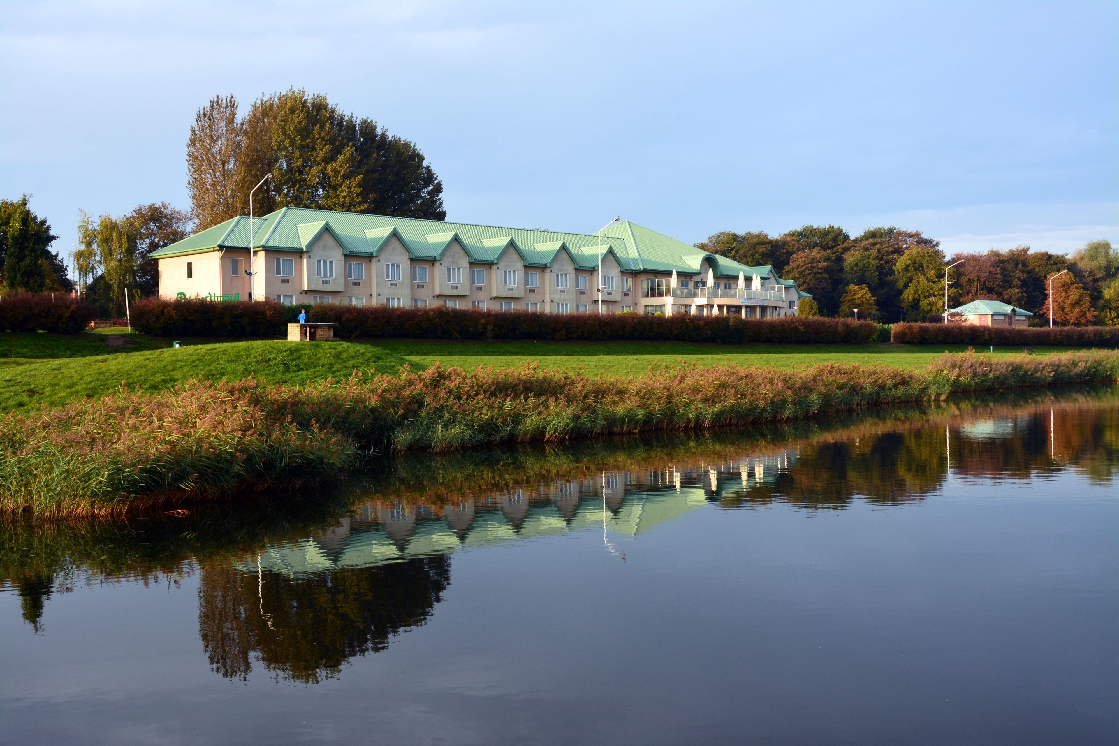 A large, multi-story building with a green roof, situated next to a calm body of water reflecting its structure.