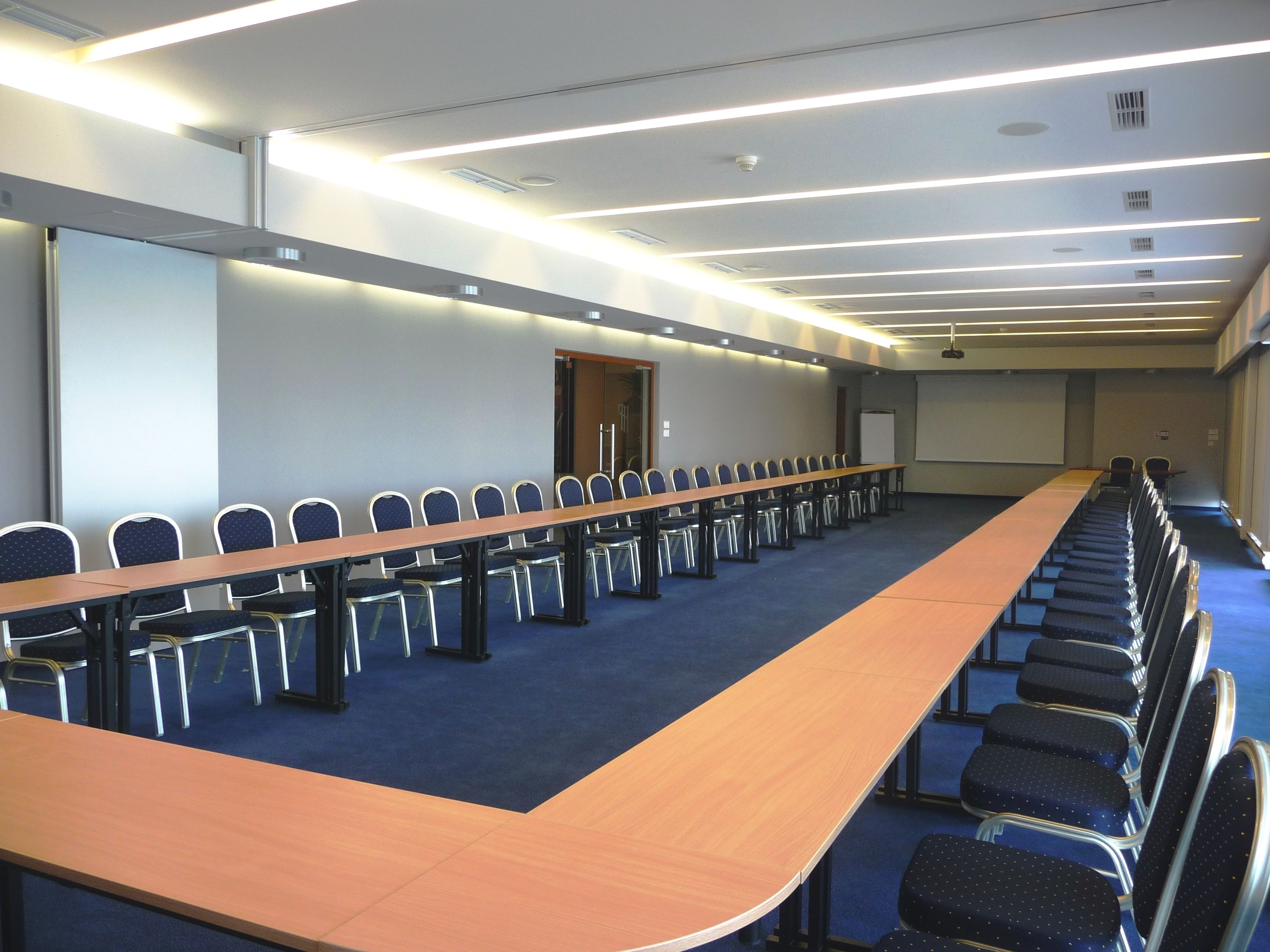 Un salón de conferencias con una larga mesa y filas de sillas, equipado con luces en el techo y una pantalla de proyector.