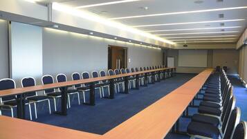 Un salón de conferencias con una larga mesa y filas de sillas, equipado con luces en el techo y una pantalla de proyector.