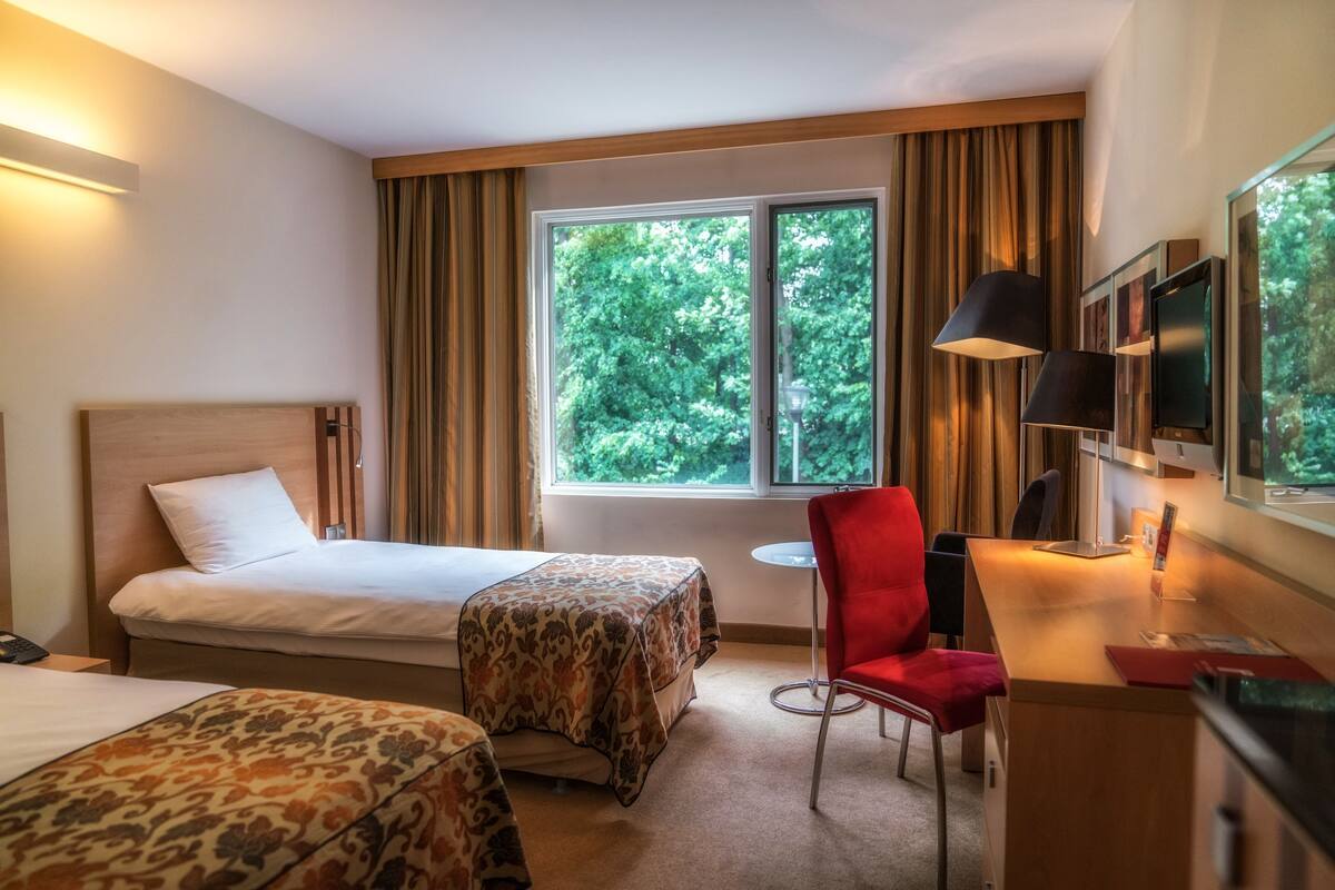 A hotel room with two beds, a desk with a chair, a TV, and a window with curtains.