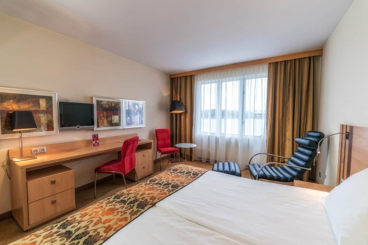 A hotel room with a bed, desk, chair, and a large window with curtains.