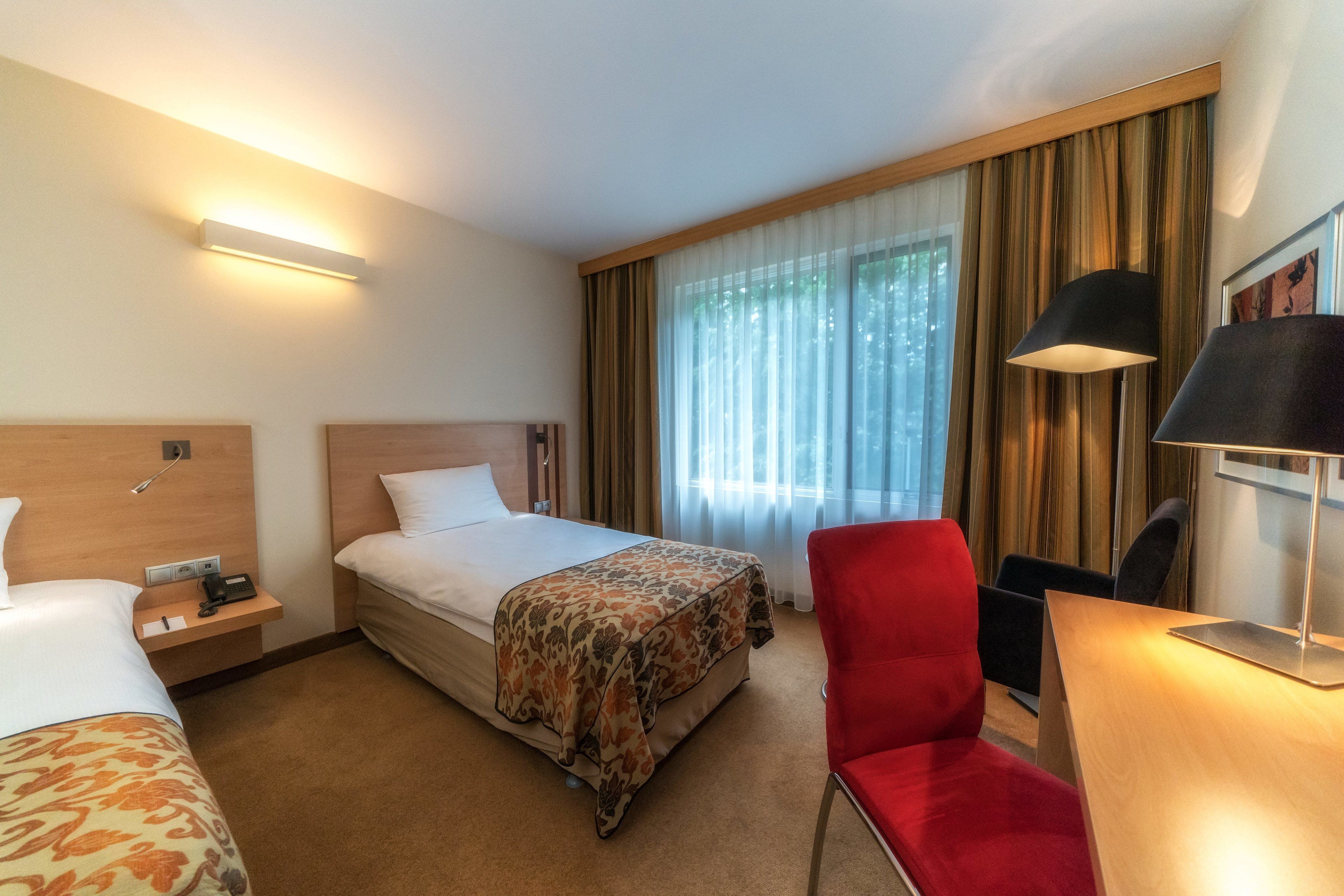A hotel room with a bed, a desk, a chair, a nightstand, and a lamp.