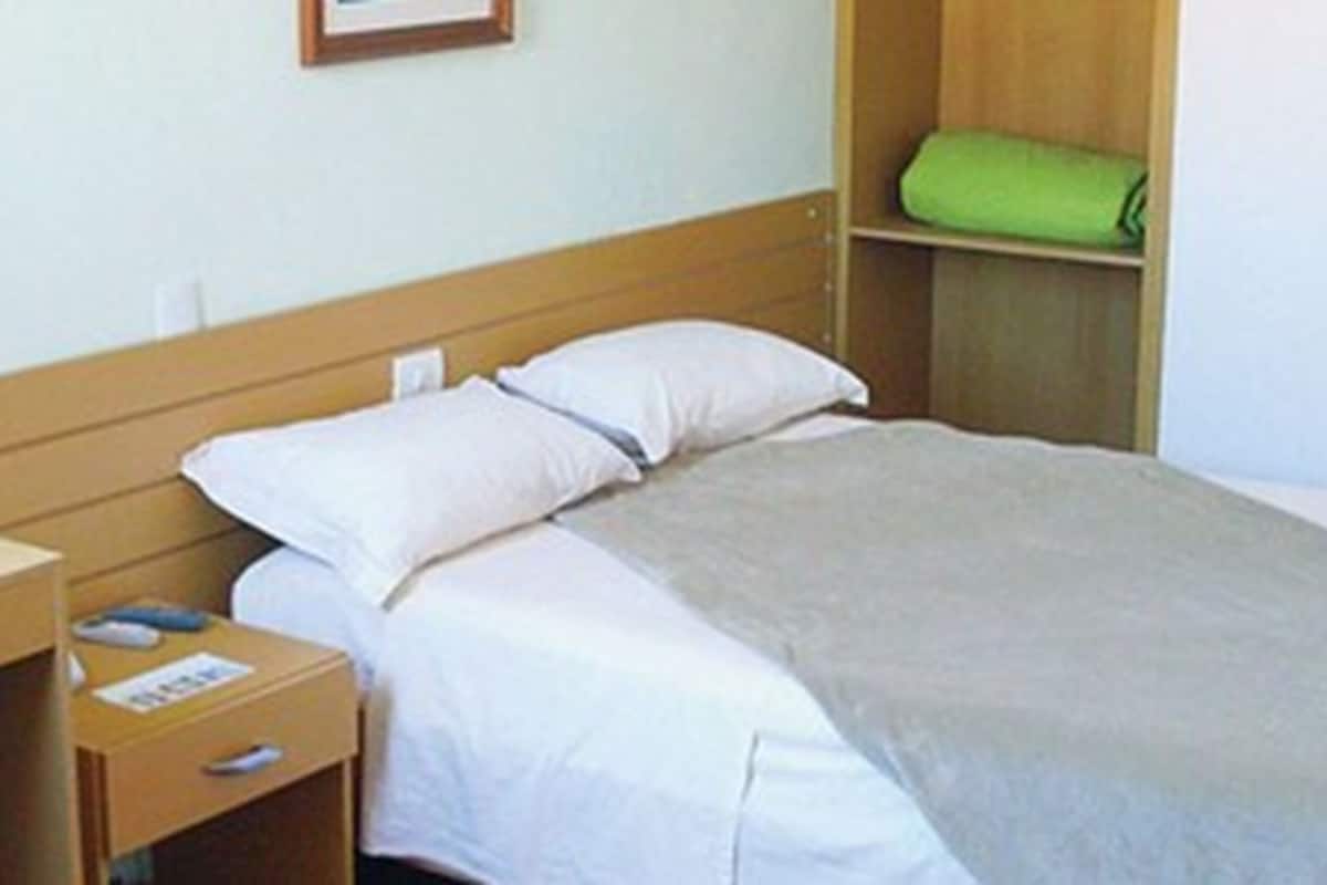 A single bed with a wooden headboard, a bedside table with a lamp, and a green towel on a shelf.