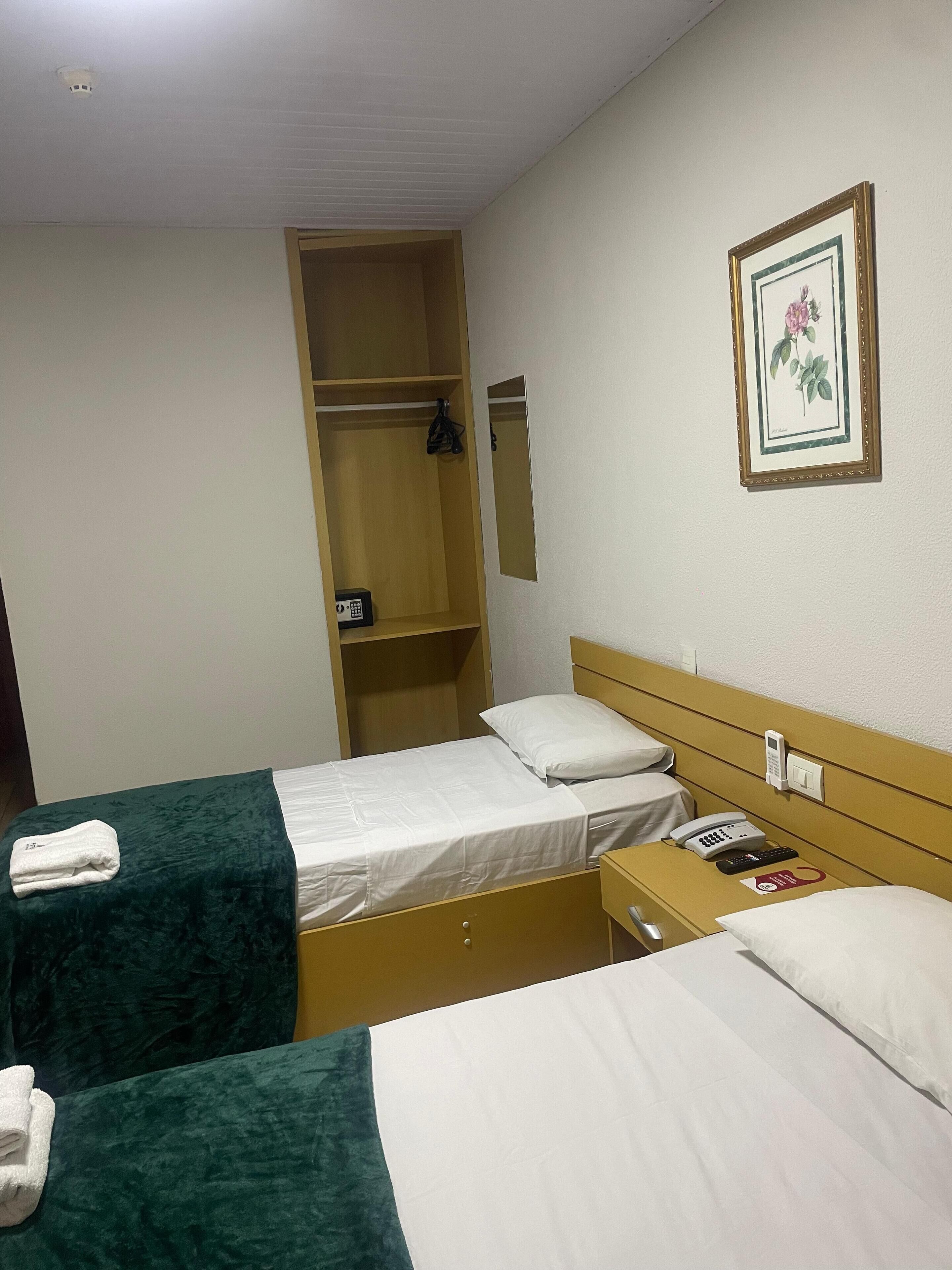 Ein Hotelzimmer mit zwei Betten, einem Kleiderschrank, einem Nachttisch mit Telefon und einem gerahmten Bild an der Wand.