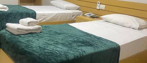 A hotel room with two single beds, each with a green bedspread and white pillows. There is a wooden wardrobe with a hanging rack, a bedside table with a phone, and a framed picture on the wall.