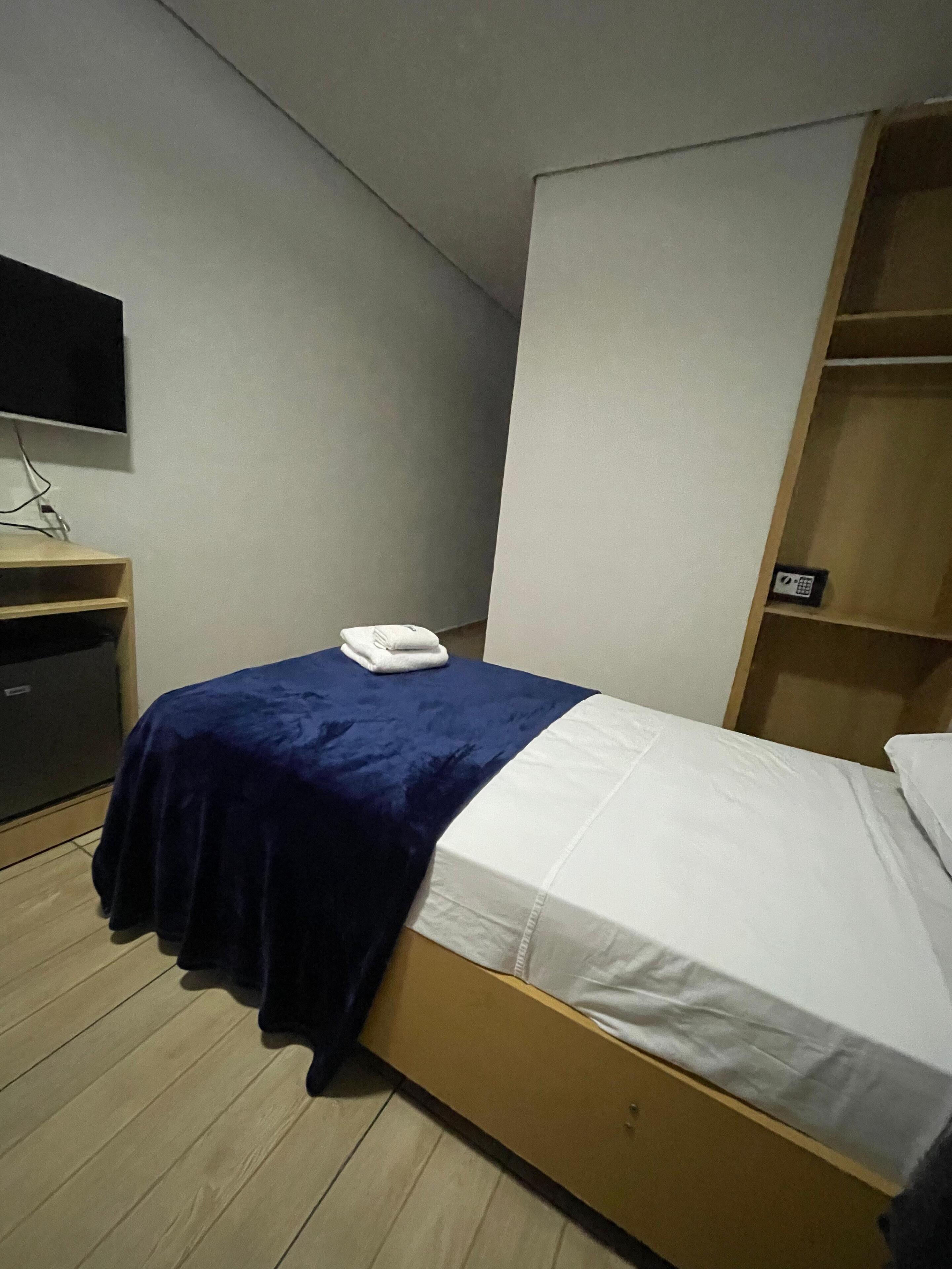 Quarto pequeno com uma cama de solteiro, chão de madeira, escrivaninha com televisão de tela plana, parede branca e prateleira com telefone.