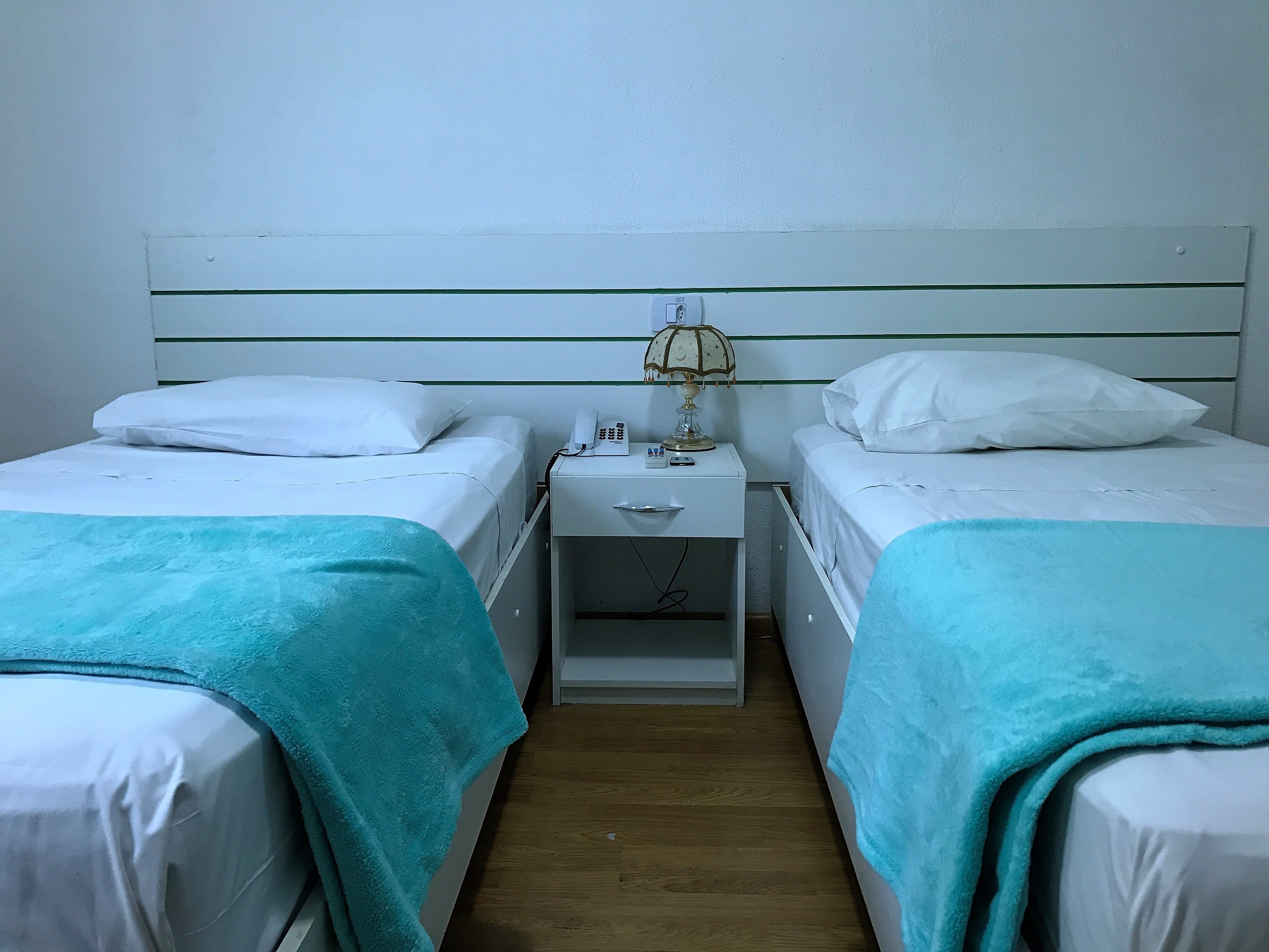 Quarto com duas camas de solteiro, cada uma com travesseiro branco e cobertor verde-água. Apresenta uma mesa de cabeceira com luminária, telefone e uma pequena gaveta. O quarto tem um design simples e clean, com piso de madeira.