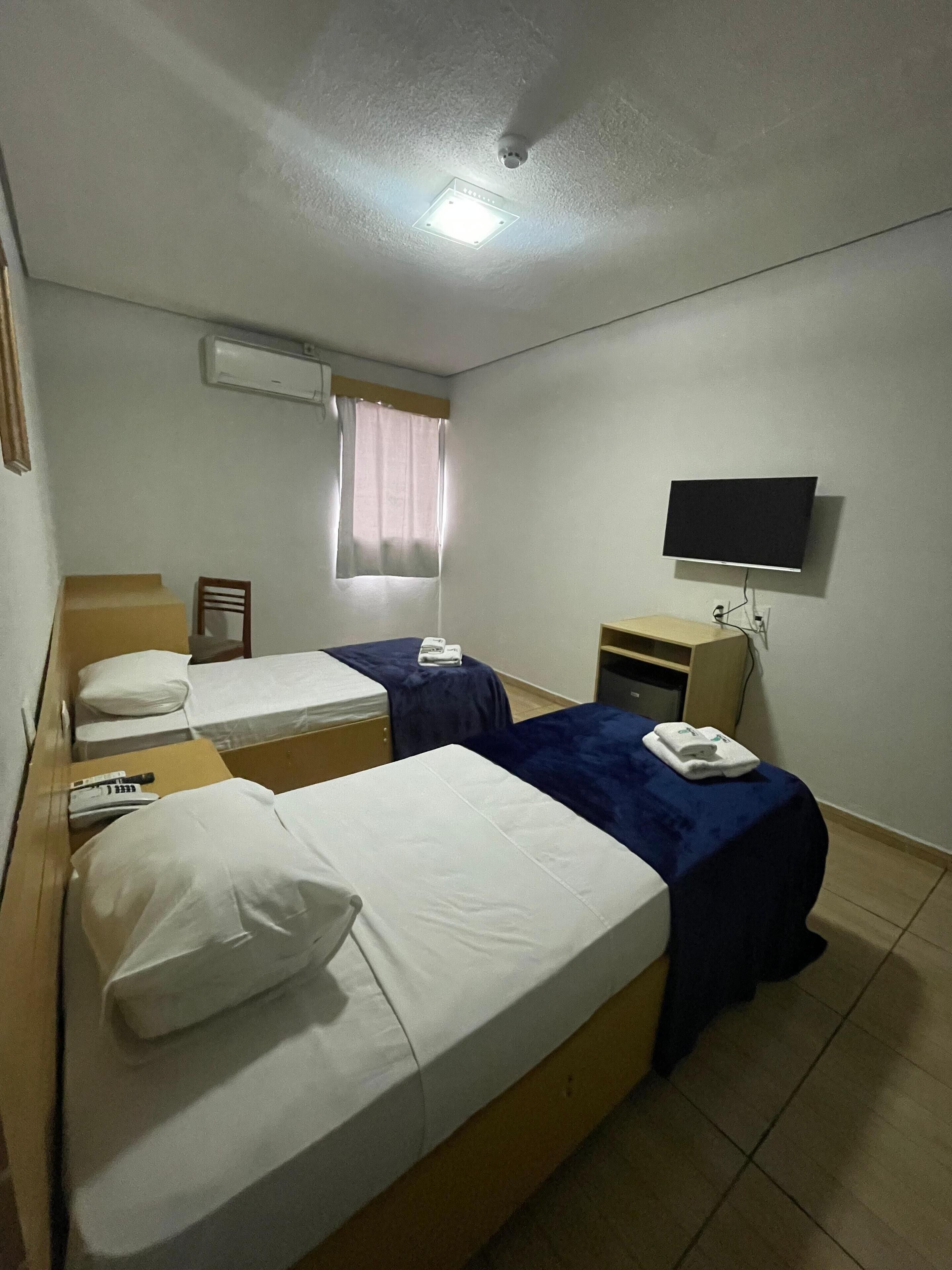 A hotel room with two beds, a TV, an air conditioning unit, and a window with curtains.