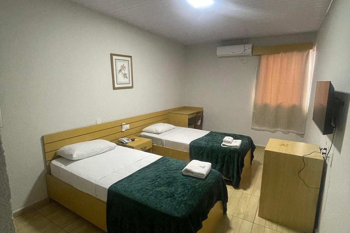 A hotel room with two beds, a desk, a chair, a TV, and an air conditioning unit.