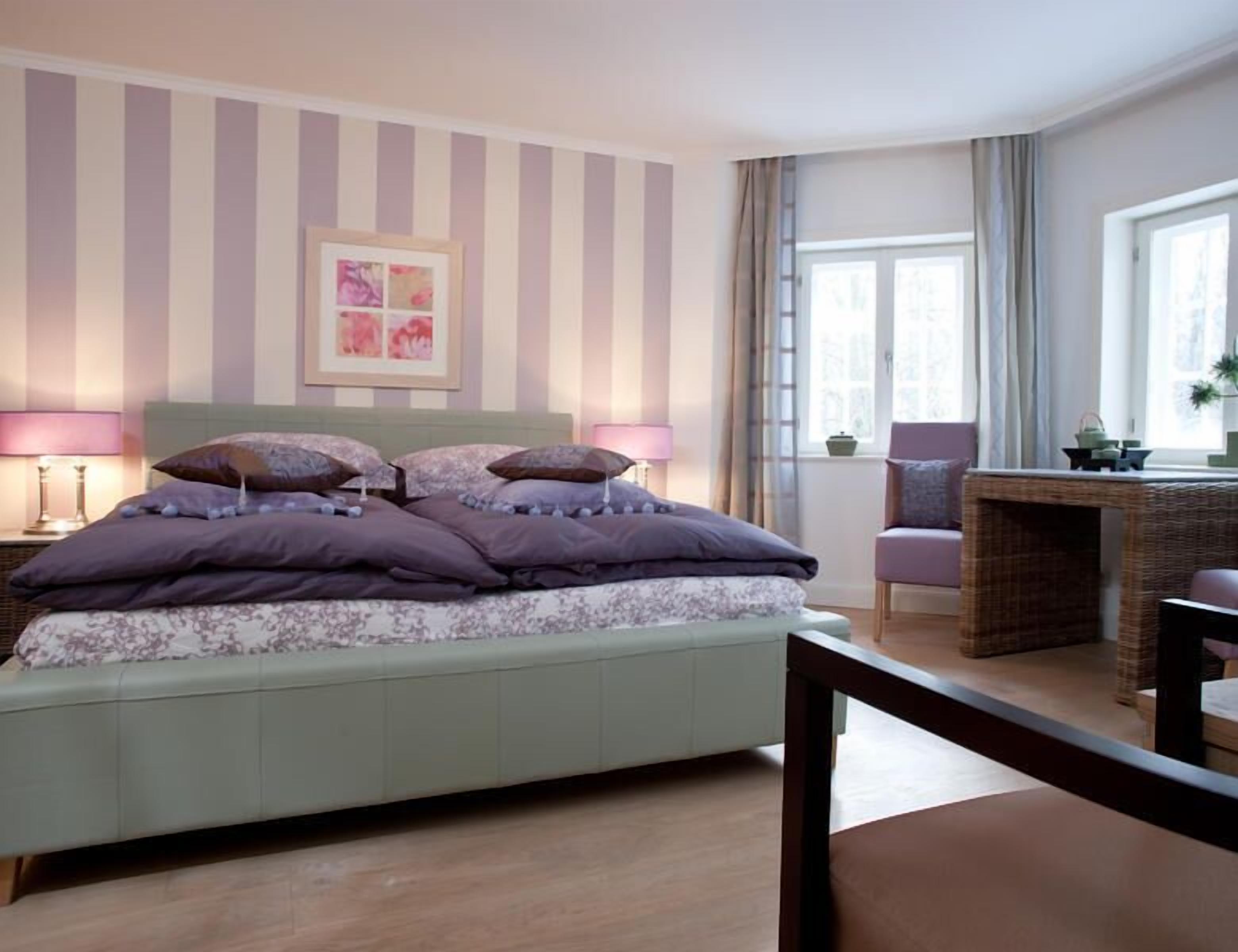 Un dormitorio con una cama, dos lámparas de noche, una obra de arte enmarcada, un ventanal con cortinas y una silla de mimbre.