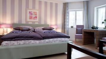 Un dormitorio con una cama, dos lámparas de noche, una obra de arte enmarcada, un ventanal con cortinas y una silla de mimbre.