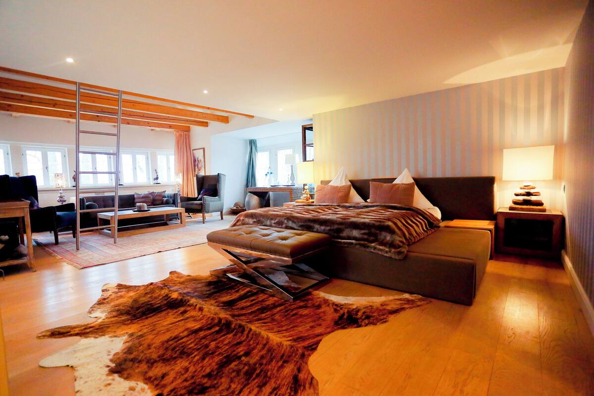 A modern bedroom with a large bed, a wooden ladder, a leather ottoman, and a fur rug.