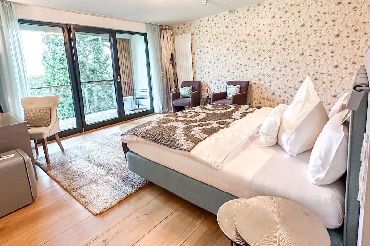 A modern bedroom with a large bed, two chairs, a small table, and a view of the outdoors through a sliding glass door.