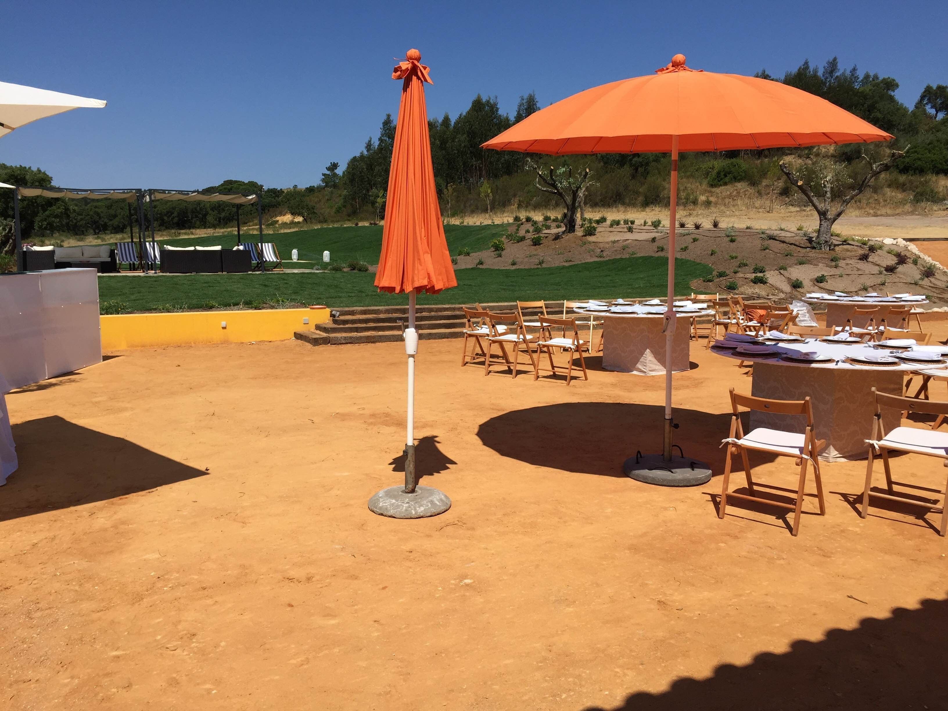 An outdoor dining area with tables and chairs, a large orange umbrella, and a view of a landscaped garden.