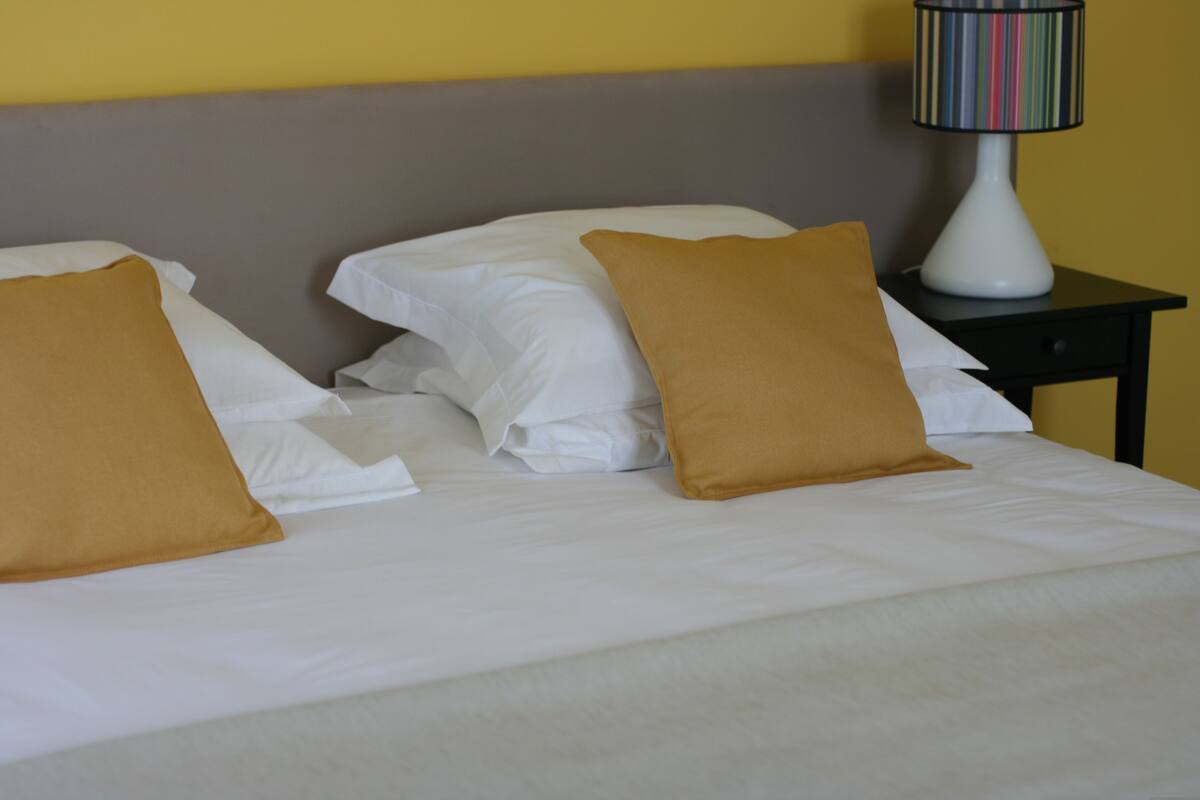 Una cama bien hecha con sábanas blancas y dos almohadas marrones, una lámpara a rayas sobre una mesita de noche, y una pared amarilla al fondo.