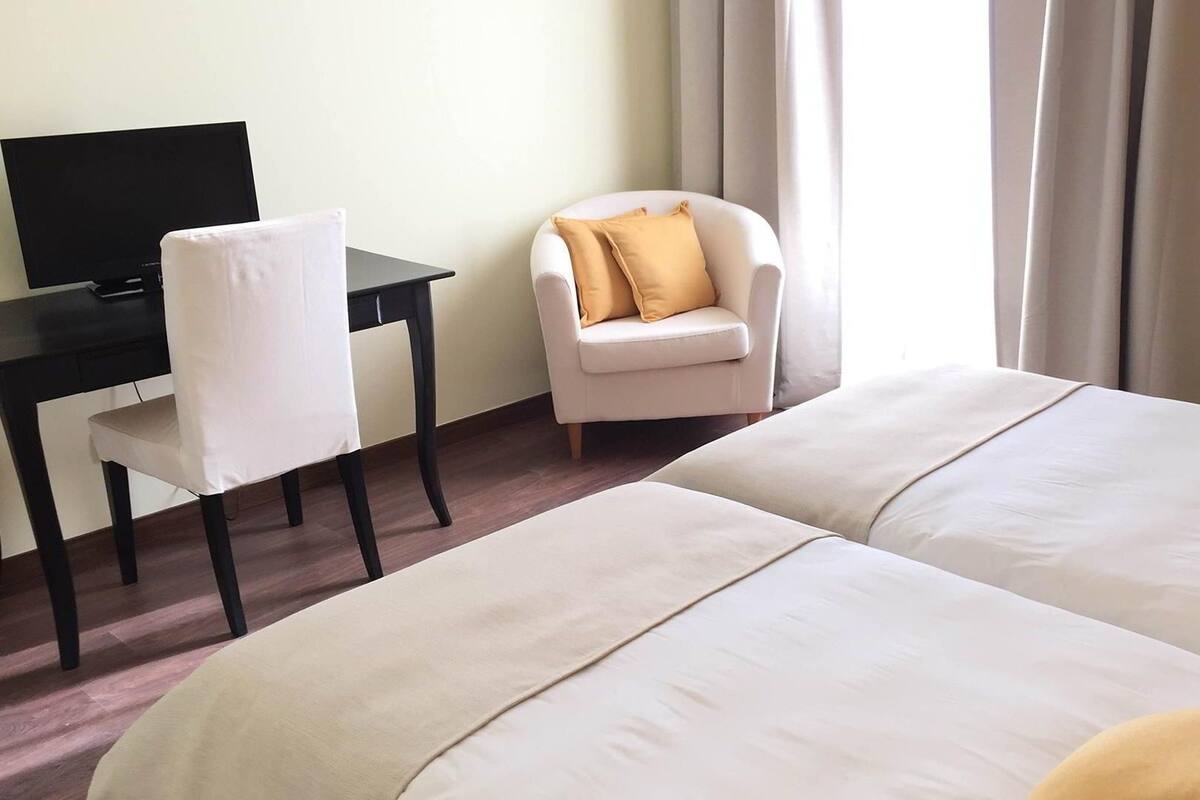 Habitación de hotel con dos camas, un escritorio con computadora, una silla y un ventanal con cortinas.