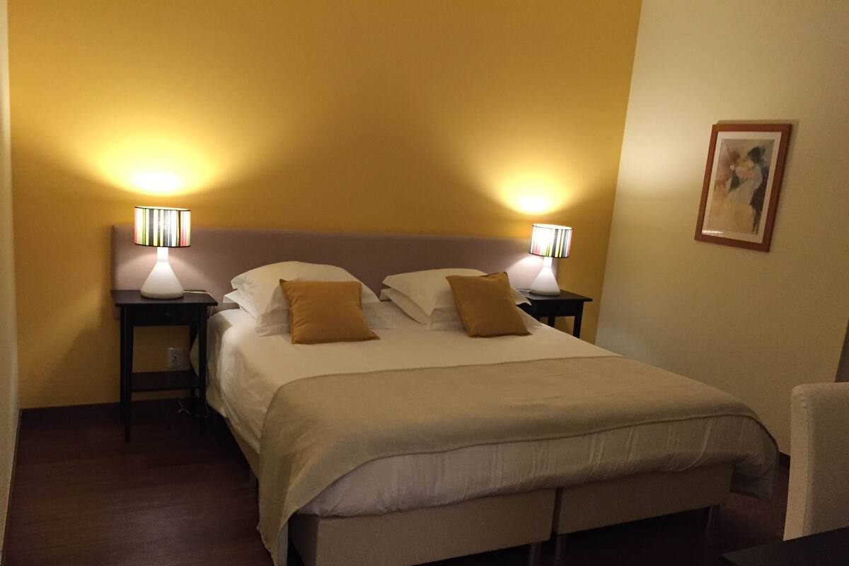 Habitación de hotel con cama, dos mesitas de noche con lámparas, una silla e imagen enmarcada en la pared.