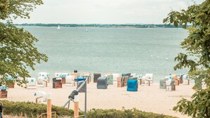 Ein Strand mit mehreren Reihen nummerierter Liegen und Sonnenschirmen, ein sandiger Uferstreifen und ein ruhiges Meer mit Segelbooten in der Ferne.