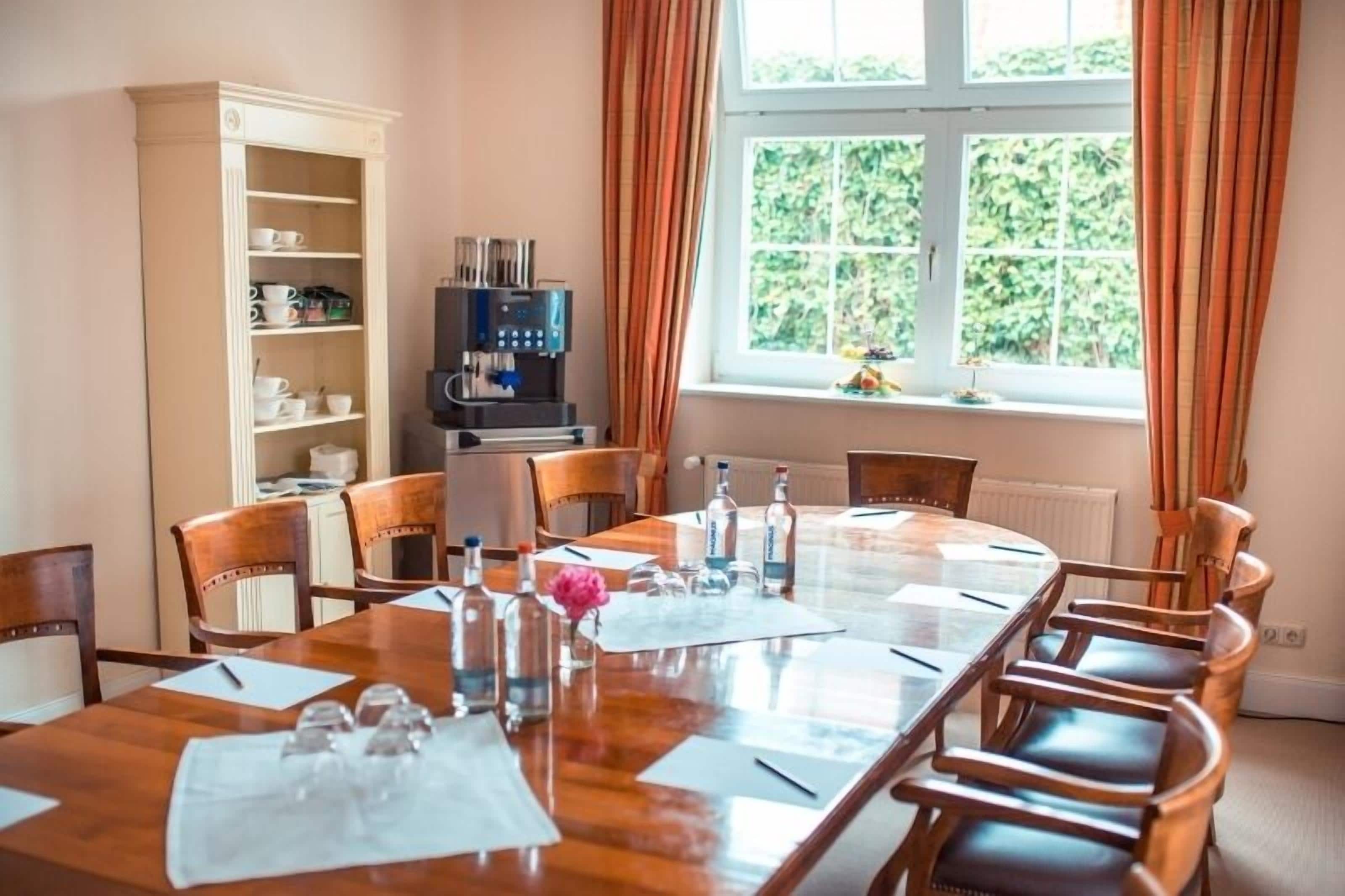 Een vergaderruimte met een grote tafel, stoelen, een koffieapparaat en een boekenplank.