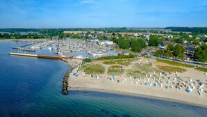 Eine Vogelperspektive auf einen Yachthafen mit Booten, einen Sandstrand mit Strandkörben und ein Wohngebiet im Hintergrund.
