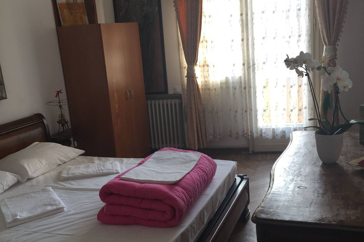 A neatly made bed with a pink blanket, a wooden wardrobe, a radiator, and a window with curtains.