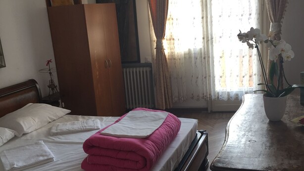 A neatly made bed with a pink blanket, a wooden wardrobe, a radiator, and a window with curtains. - Ca' Barbaria (Venice)
