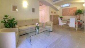 A modern living room with a sofa, coffee table, and chairs, a dining area with a table and chairs, and a kitchen in the background. - Palazzo Ruschioni Boutique Hotel (Camerano)