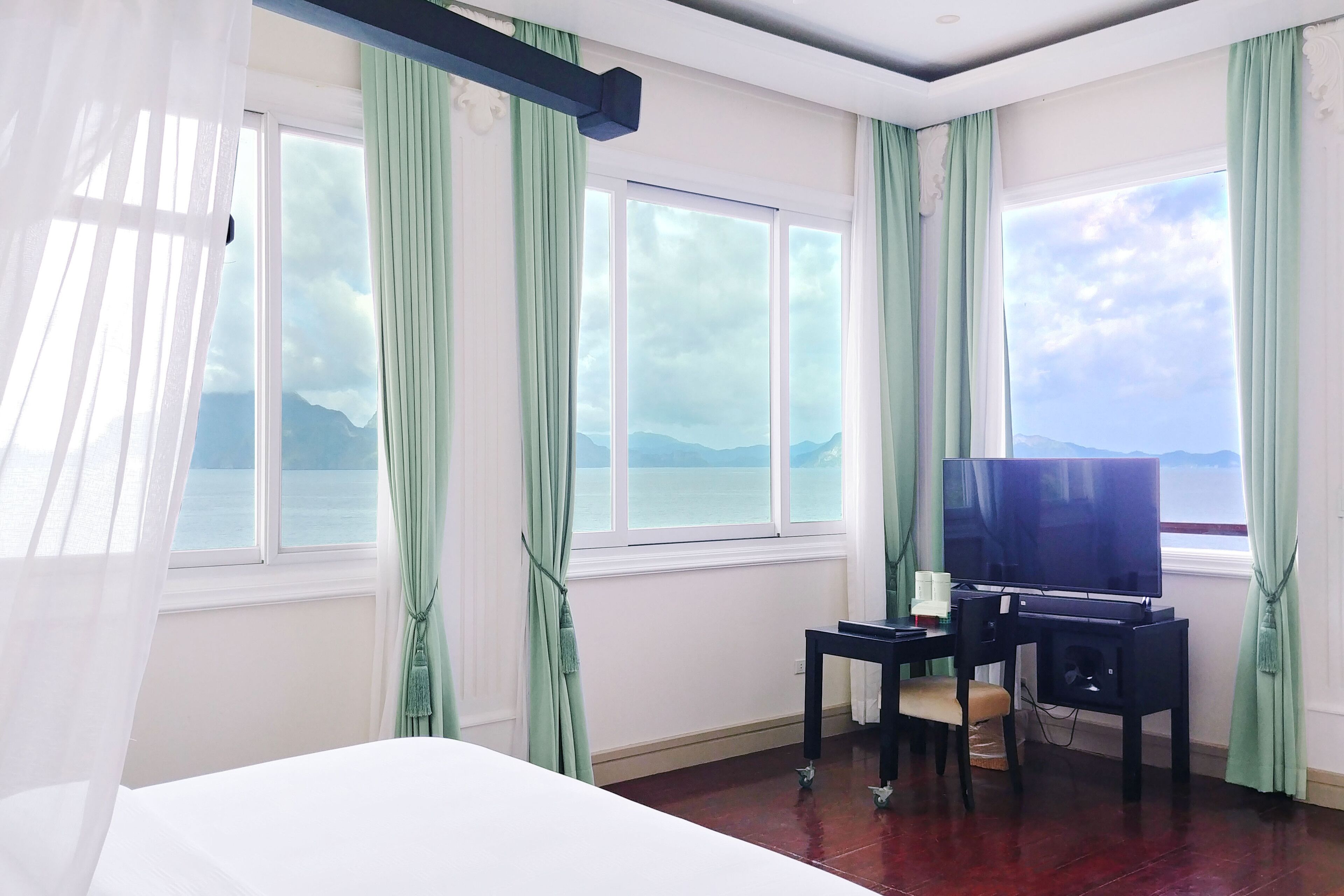 A hotel room with a bed, a desk with a television, and large windows with curtains.